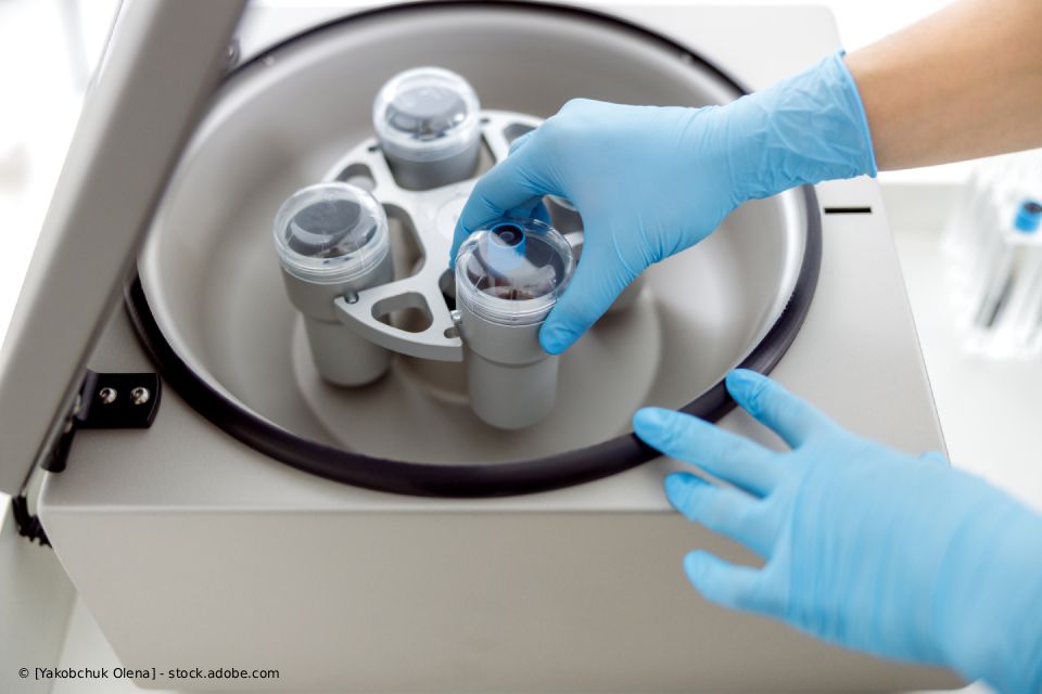 Behandschuhte Hände stellen ein Reagenzglas in eine Zentrifuge. Weiße Laborgeräte mit Reagenzgläsern auf einem rotierenden Apparat.