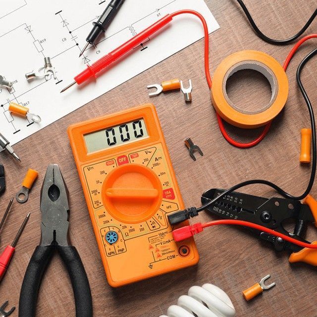 Multímetro naranja con herramientas y componentes eléctricos sobre una superficie de madera.