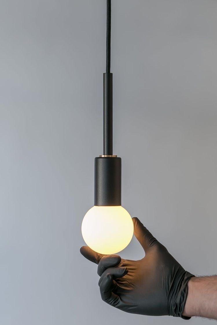 Mano en guante negro que sostiene una bombilla redonda encendida suspendida de una lámpara de techo negra.