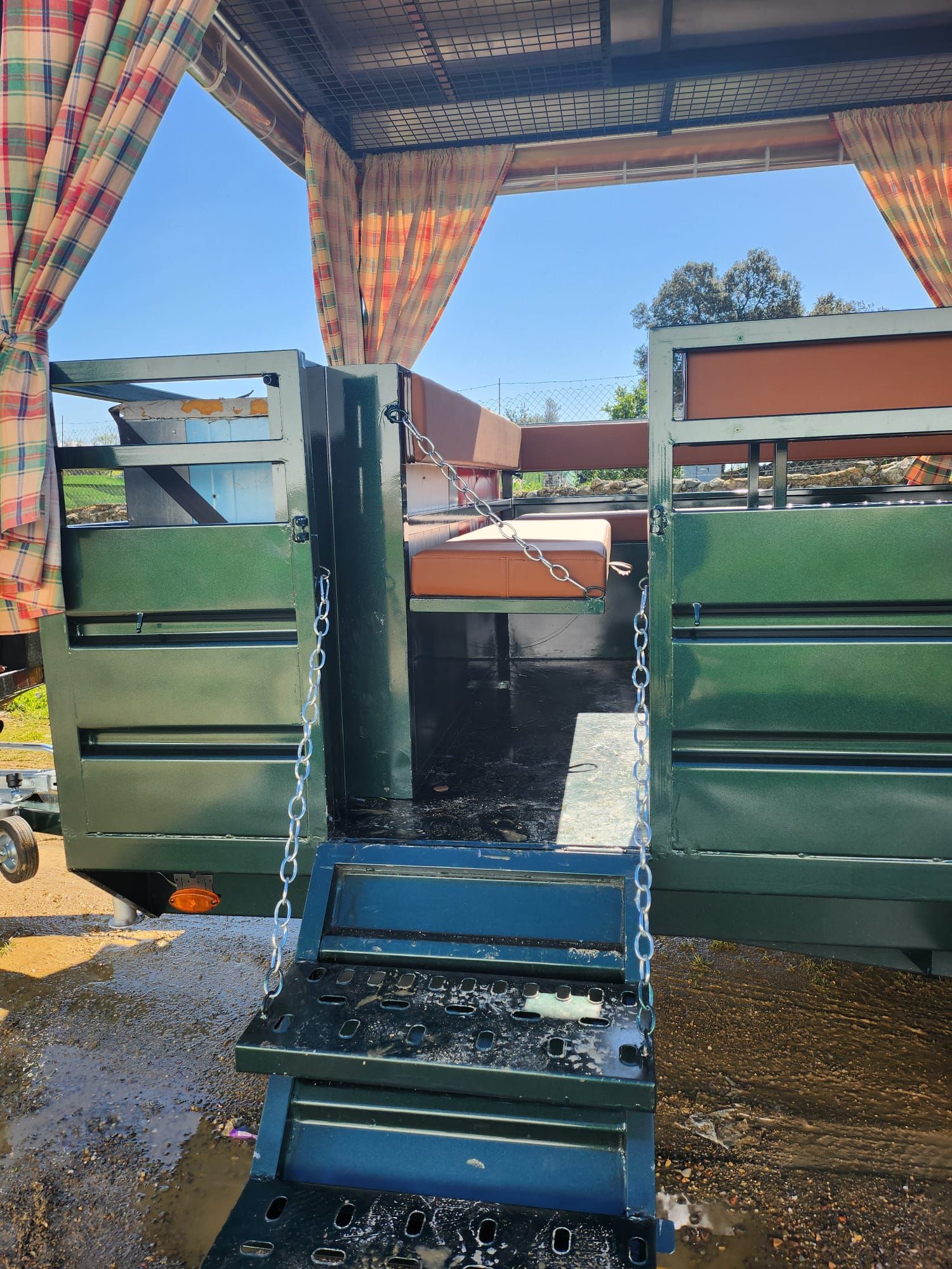 Remolque verde con puertas abiertas y escaleras bajo un toldo colorido, al aire libre en un día soleado.