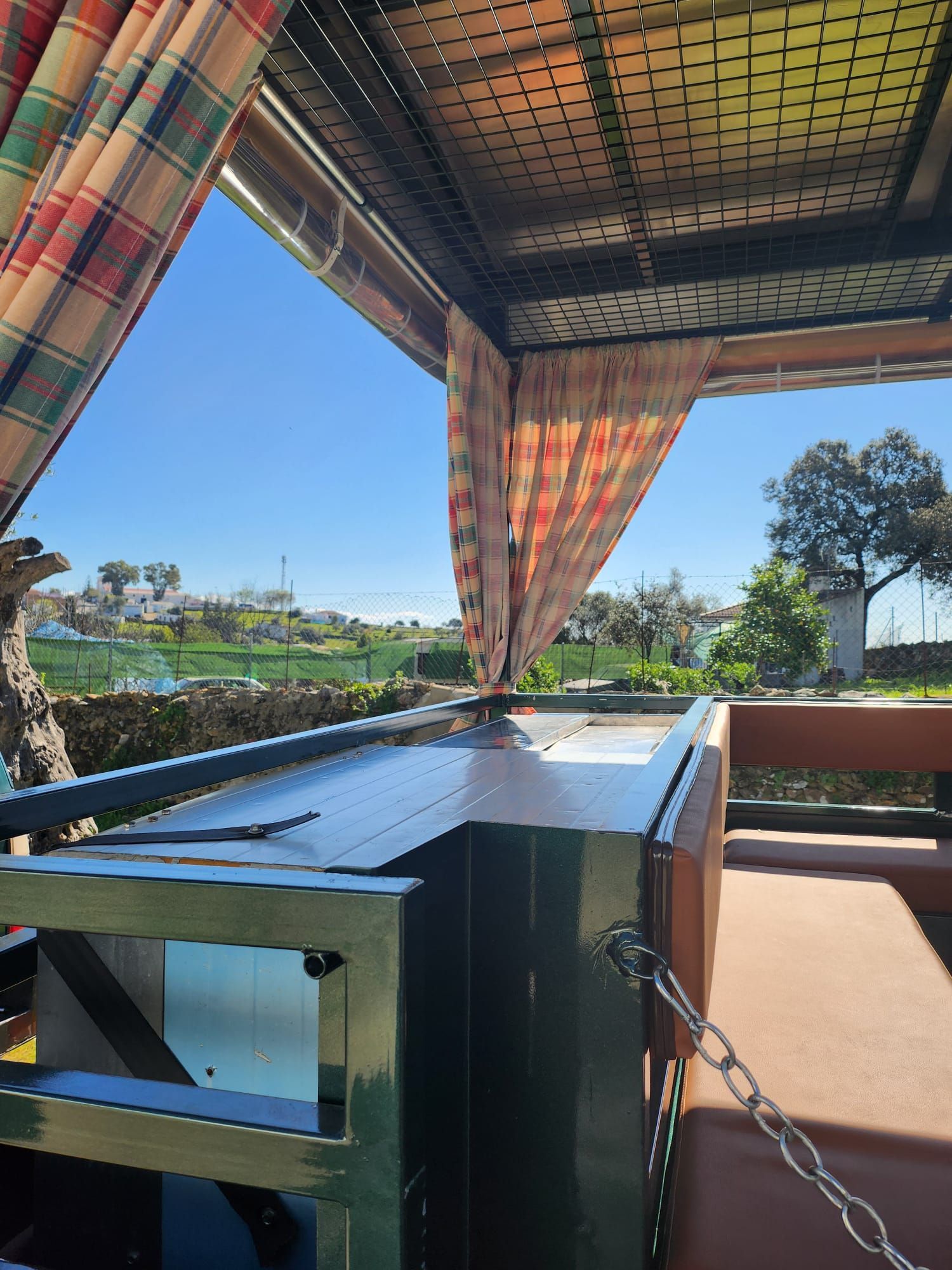 Zona de estar al aire libre con parrilla de metal verde, cortinas a cuadros, paisaje soleado.