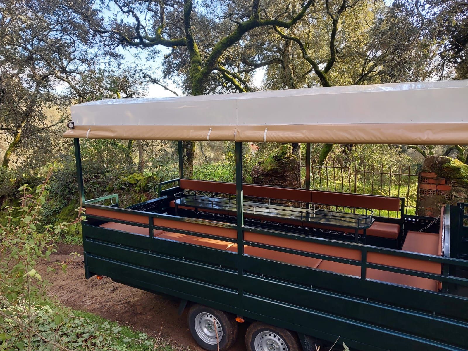 Remolque tipo safari de color verde, con bancos bajo un toldo beige, estacionado en una zona boscosa.