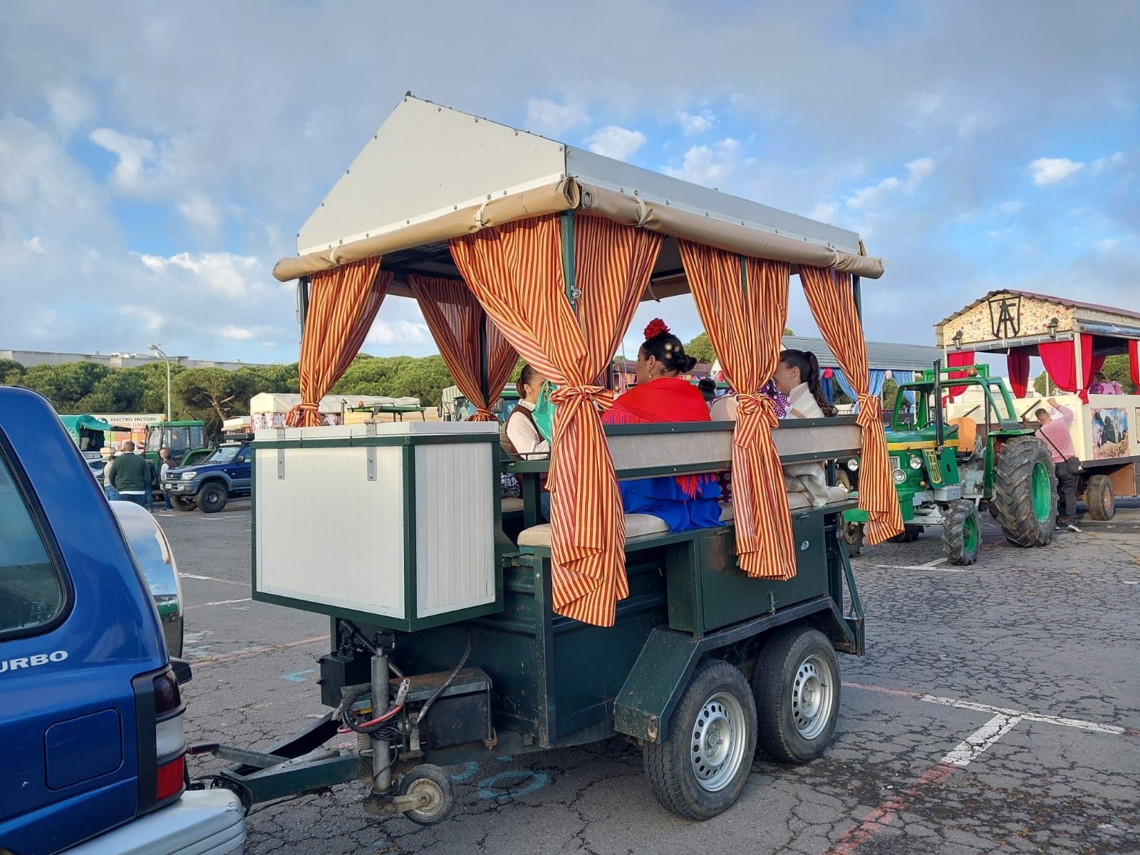 Remolque verde con toldo y cortinas naranjas, con pasajeros. En el estacionamiento.
