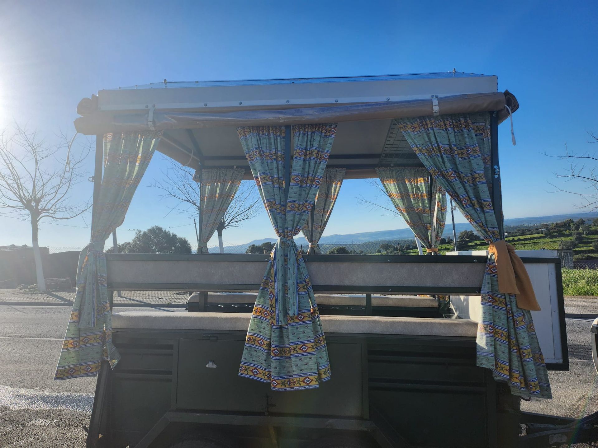 Un bar móvil, verde oliva con cortinas florales azules, bajo un cielo azul brillante.