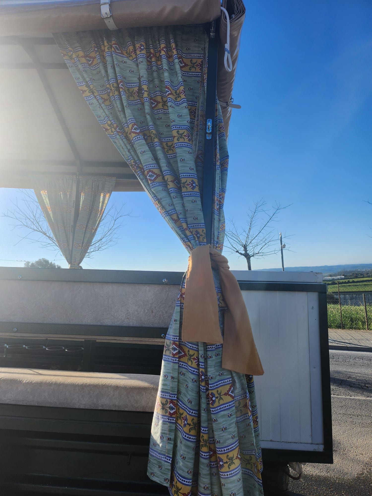 Exterior de autocaravana, cortinas estampadas, cielo azul, día soleado.