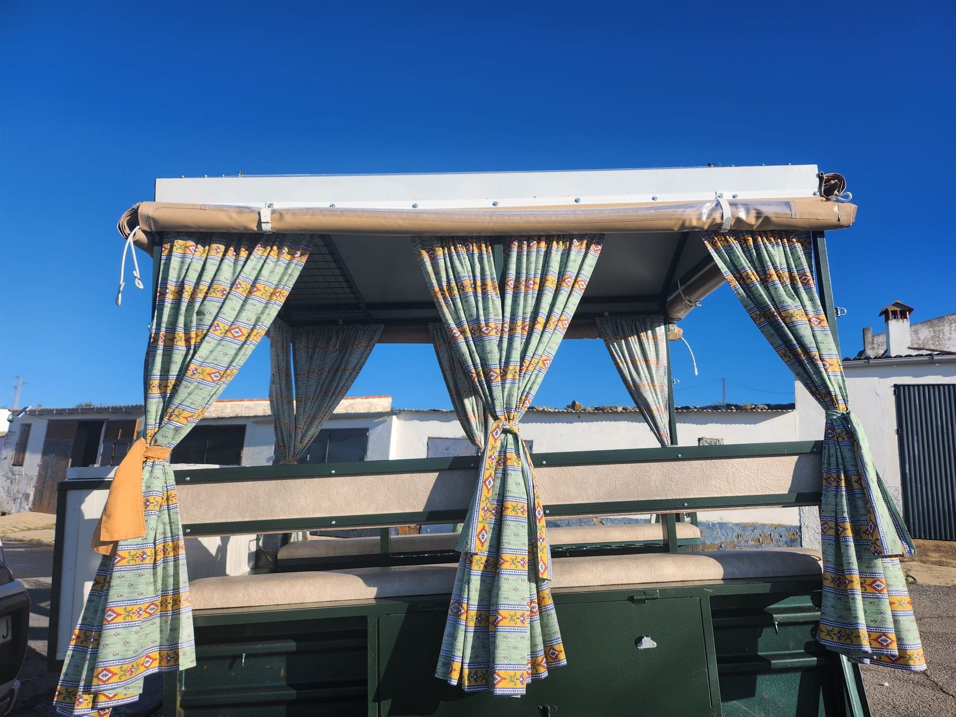 Carruaje tirado por caballos verde con cortinas estampadas bajo un cielo azul.