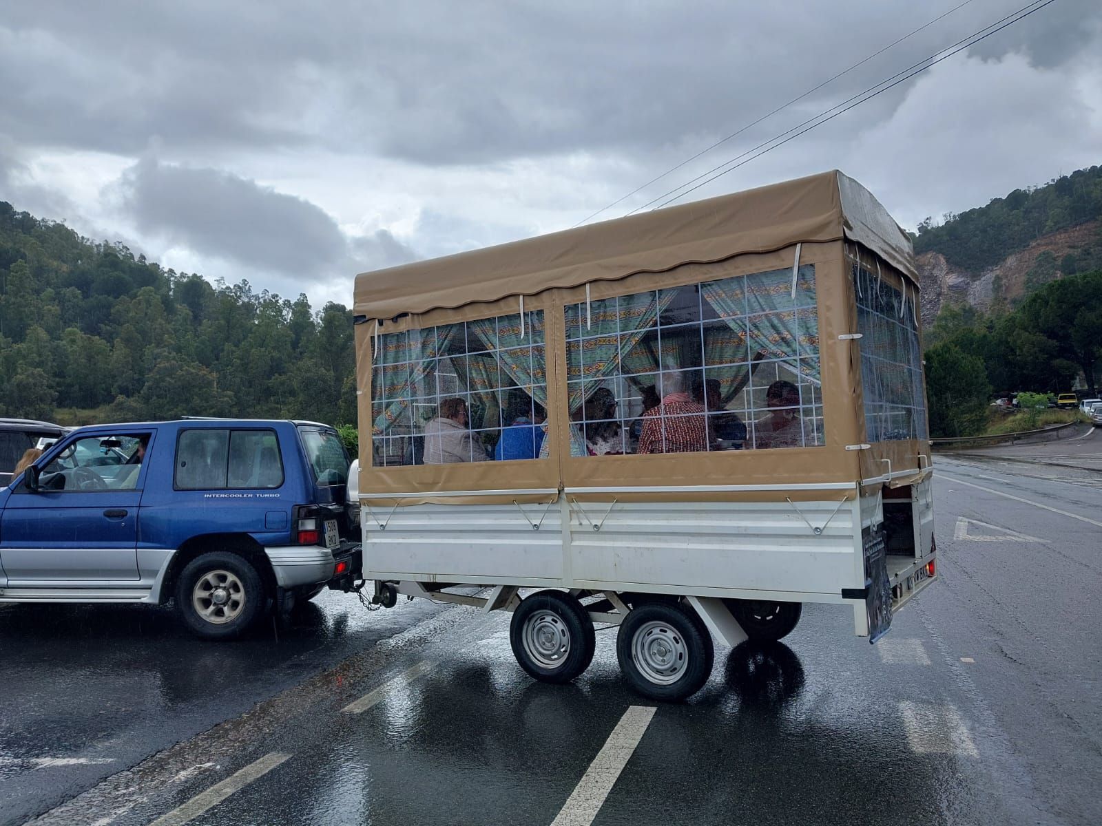 Una camioneta todoterreno azul remolcando un remolque con techo de lona color marrón, llena de gente, en una carretera mojada cerca de árboles.