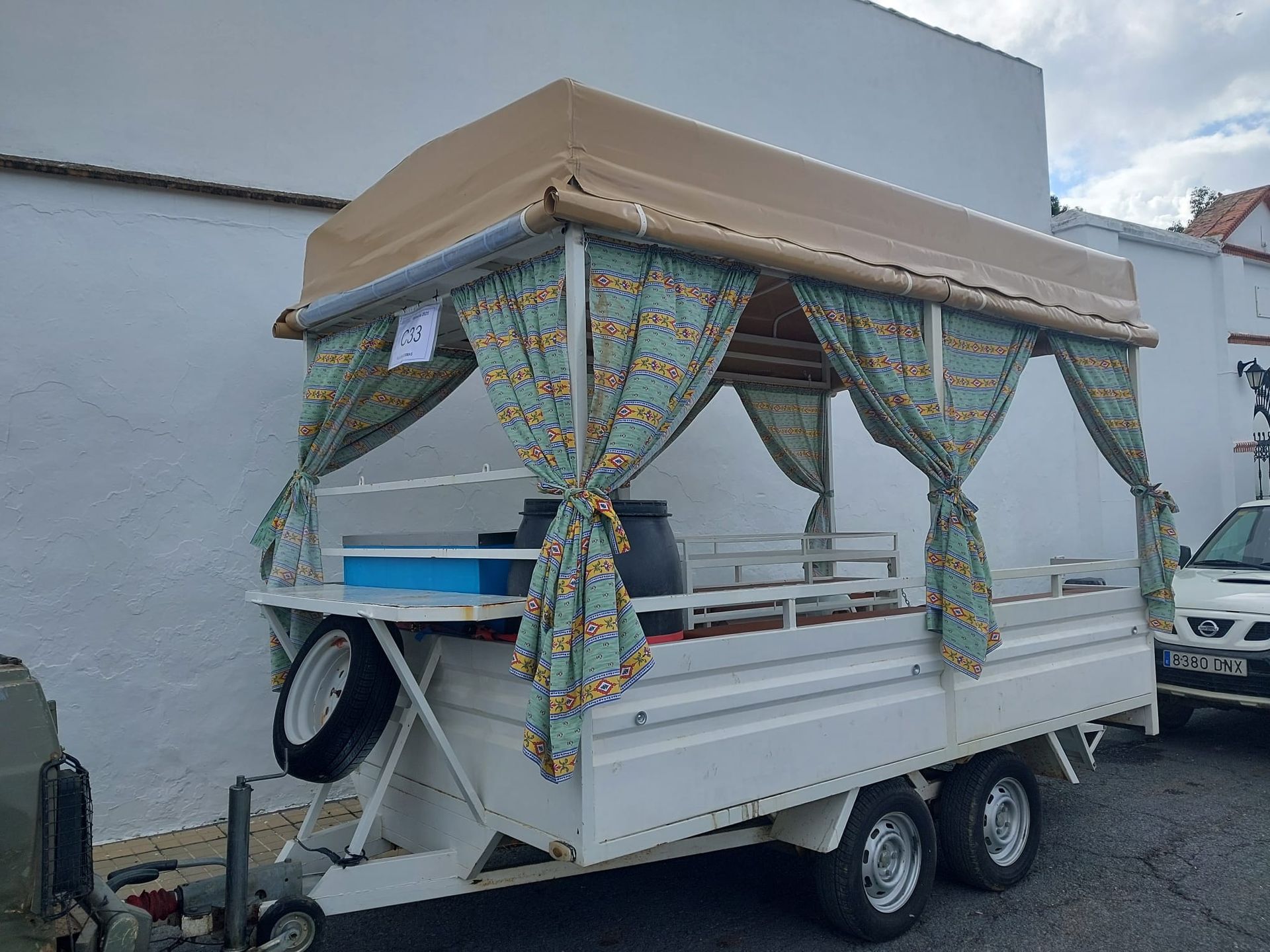 Remolque blanco con toldo y cortinas de color tostado, utilizado como puesto al aire libre, estacionado en una calle.