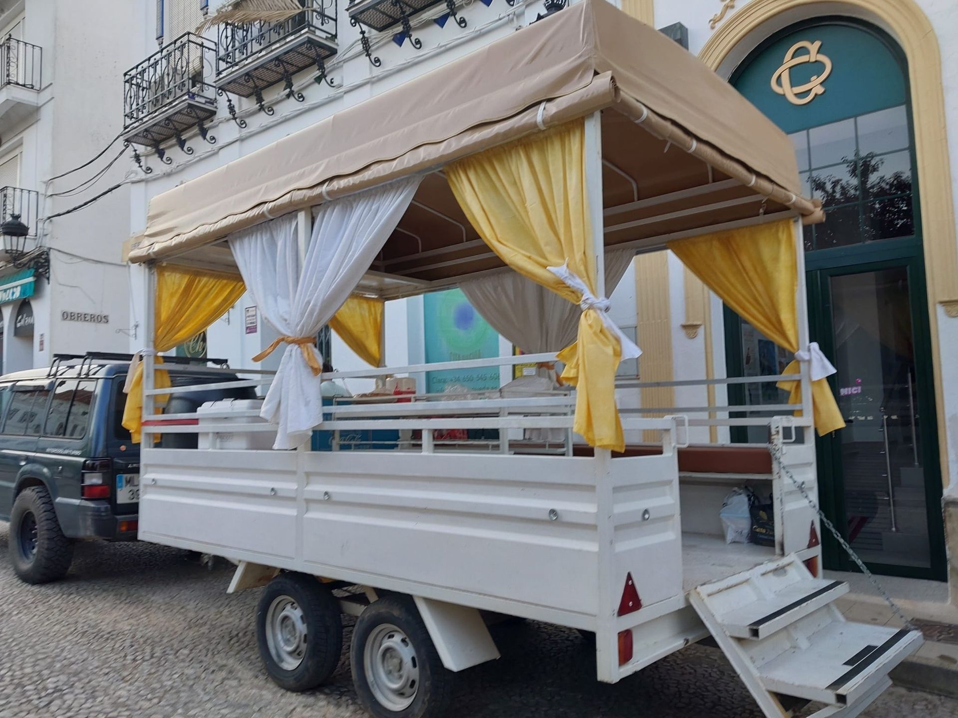 Un remolque blanco con un toldo color tostado y cortinas de color amarillo y blanco, estacionado en una calle.