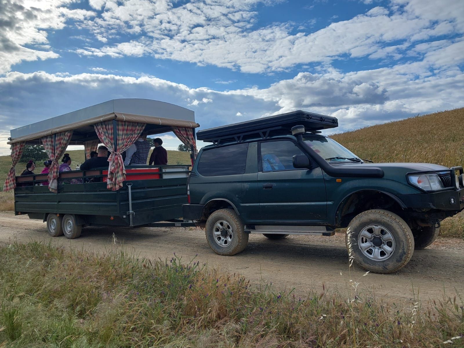 Una camioneta verde oscuro arrastra un remolque con toldo; hay personas dentro. Circula por un camino de tierra, con un fondo montañoso.