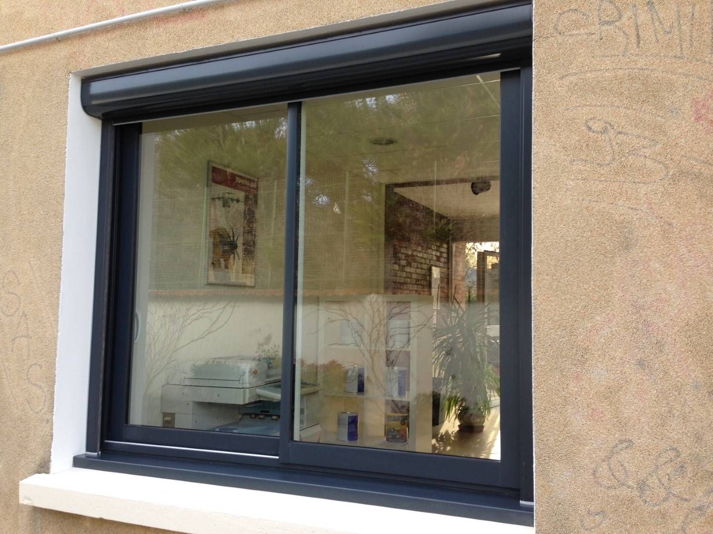 Fenêtre coulissante à cadre foncé avec un volet roulant gris foncé fermé, encastrée dans un mur en stuc beige.