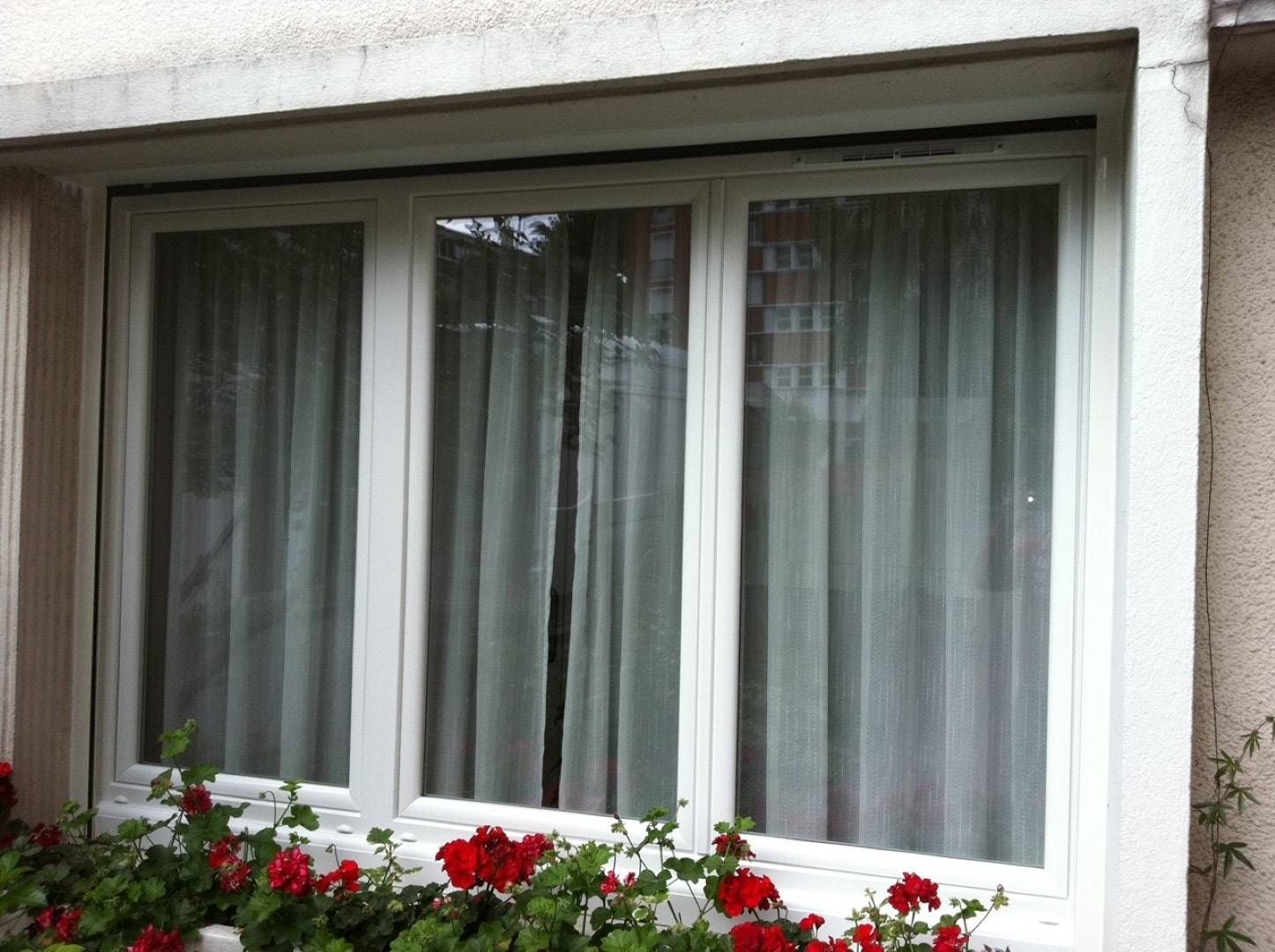 Fenêtre à cadre blanc avec rideaux transparents et fleurs rouges dans une jardinière.