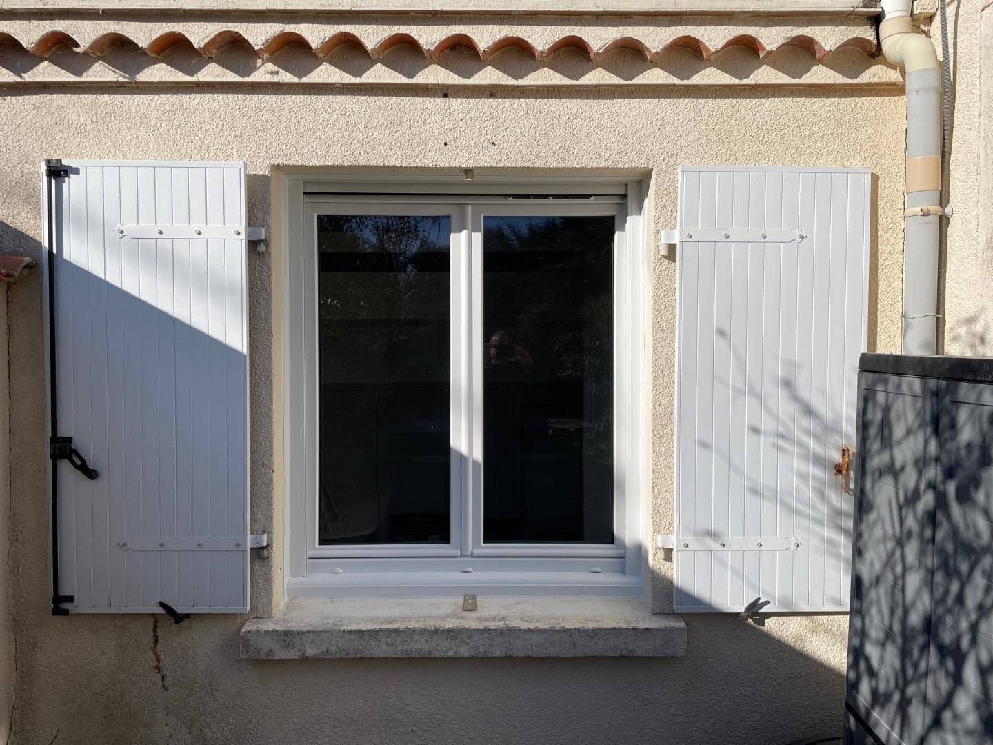 Des volets blancs encadrent une fenêtre à double vitrage sur un mur en stuc. On y trouve également une petite corniche en béton et une gouttière.