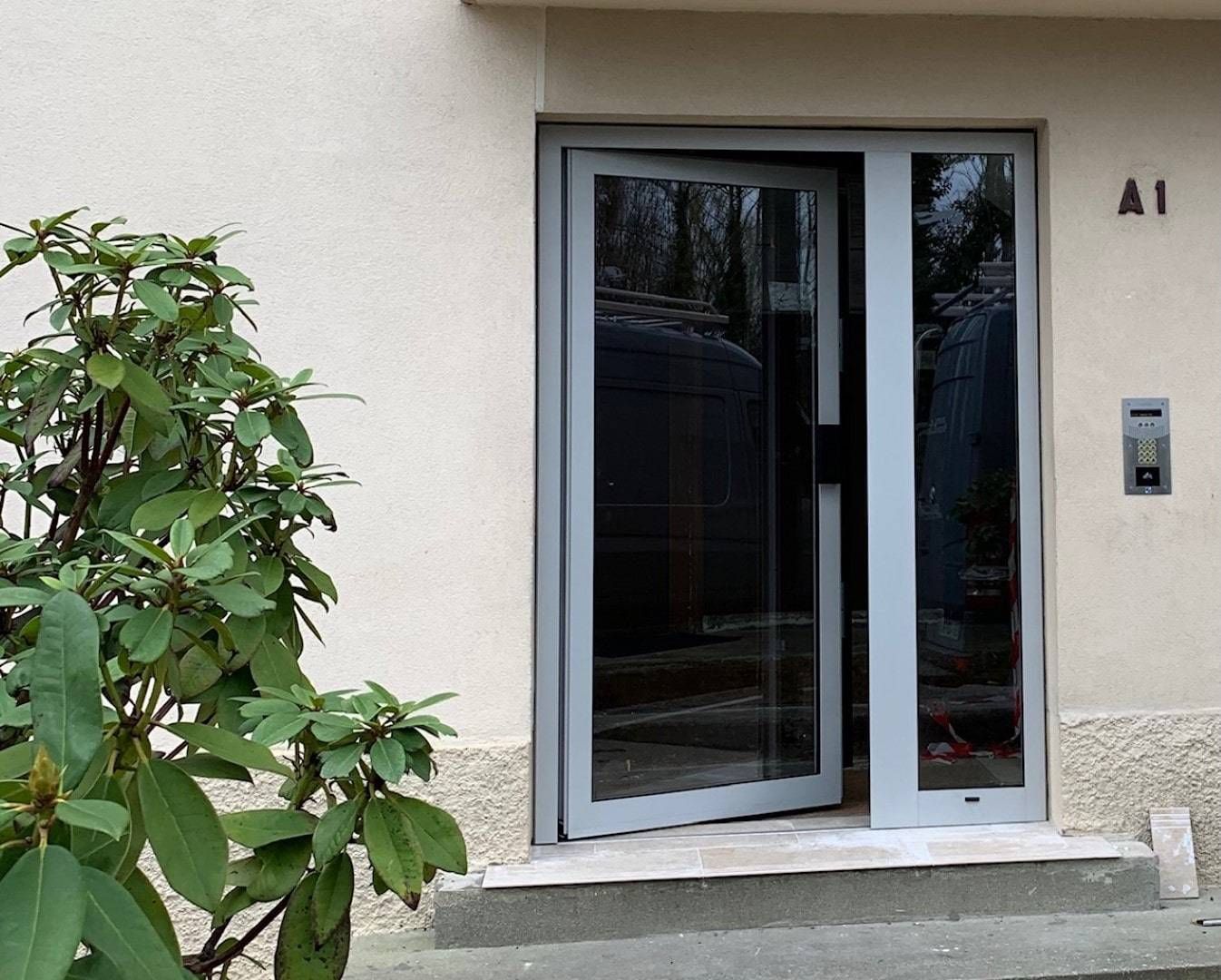 Porte d'entrée en verre gris légèrement entrouverte, à côté d'un panneau vitré fixe. « A1 » au-dessus de la porte.