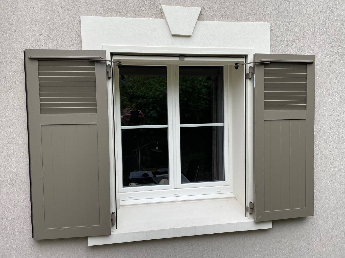 Fenêtre avec volets gris ouverts, garniture blanche sur un mur beige.