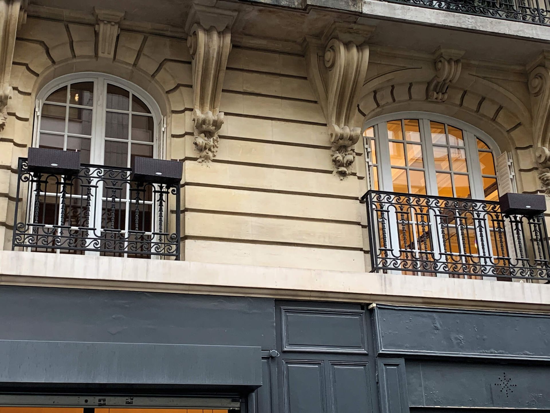 Deux fenêtres cintrées avec balcons en fer forgé sur une façade claire. Auvent gris en dessous.