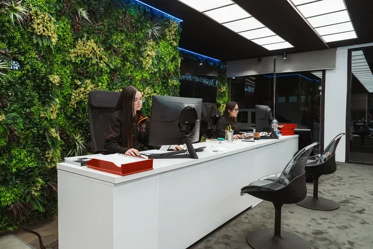 Dos personas trabajan en un largo mostrador de recepción blanco, frente a una pared vegetal verde, dentro de un espacio de oficinas moderno.