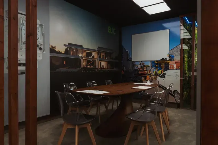 Una moderna sala de reuniones cuenta con una mesa central de madera oscura rodeada de seis sillas bajo una lámpara de techo rectangular.