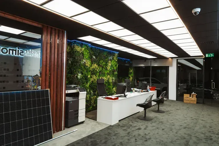 Una luminosa zona de recepción de oficina con un escritorio blanco, una pared de musgo verde, paneles solares e iluminación moderna en el techo.