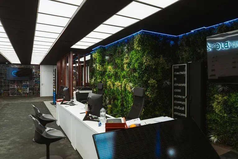 Una moderna zona de recepción con un largo escritorio blanco, sillas giratorias y una exuberante pared vegetal de color verde bajo una iluminación brillante.