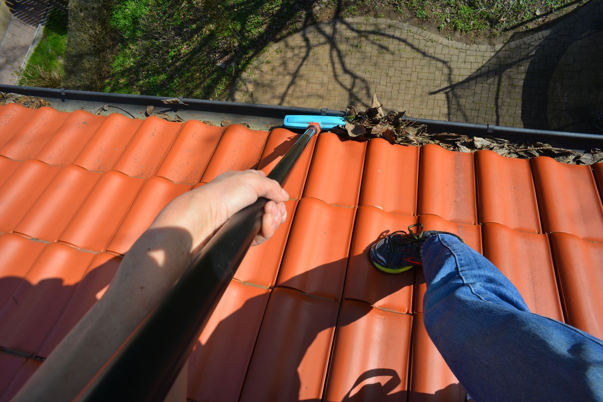Une personne sur un toit de tuiles utilise un outil pour nettoyer une gouttière.