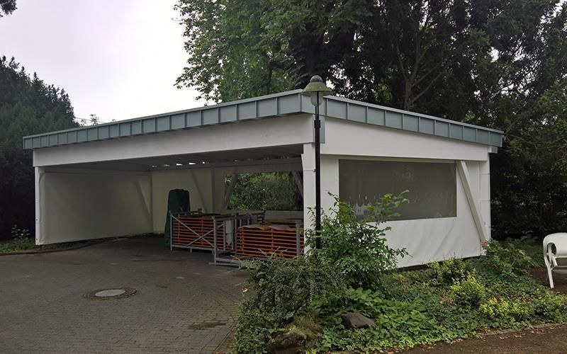 Ein weißer Carport mit blauem Dach ist von Bäumen und Büschen umgeben.