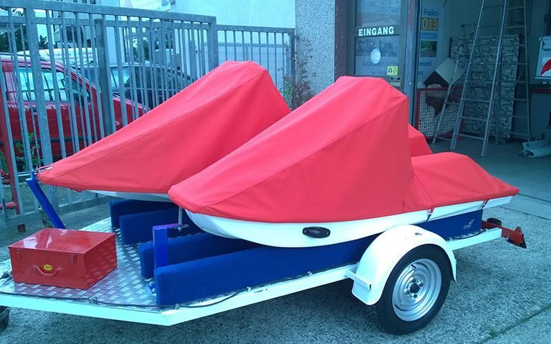 Auf einem Anhänger stehen zwei Jetskis mit roten und blauen Planen.