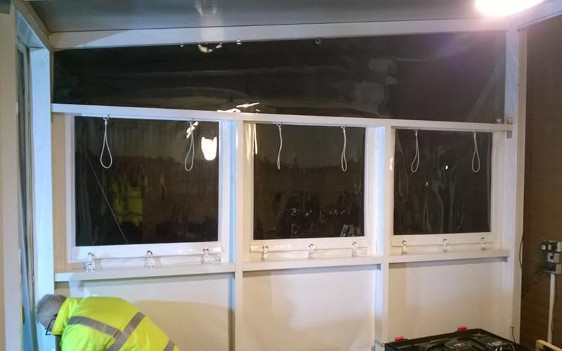 Ein Mann in einer gelben Jacke steht vor einer Wand mit drei Fenstern.