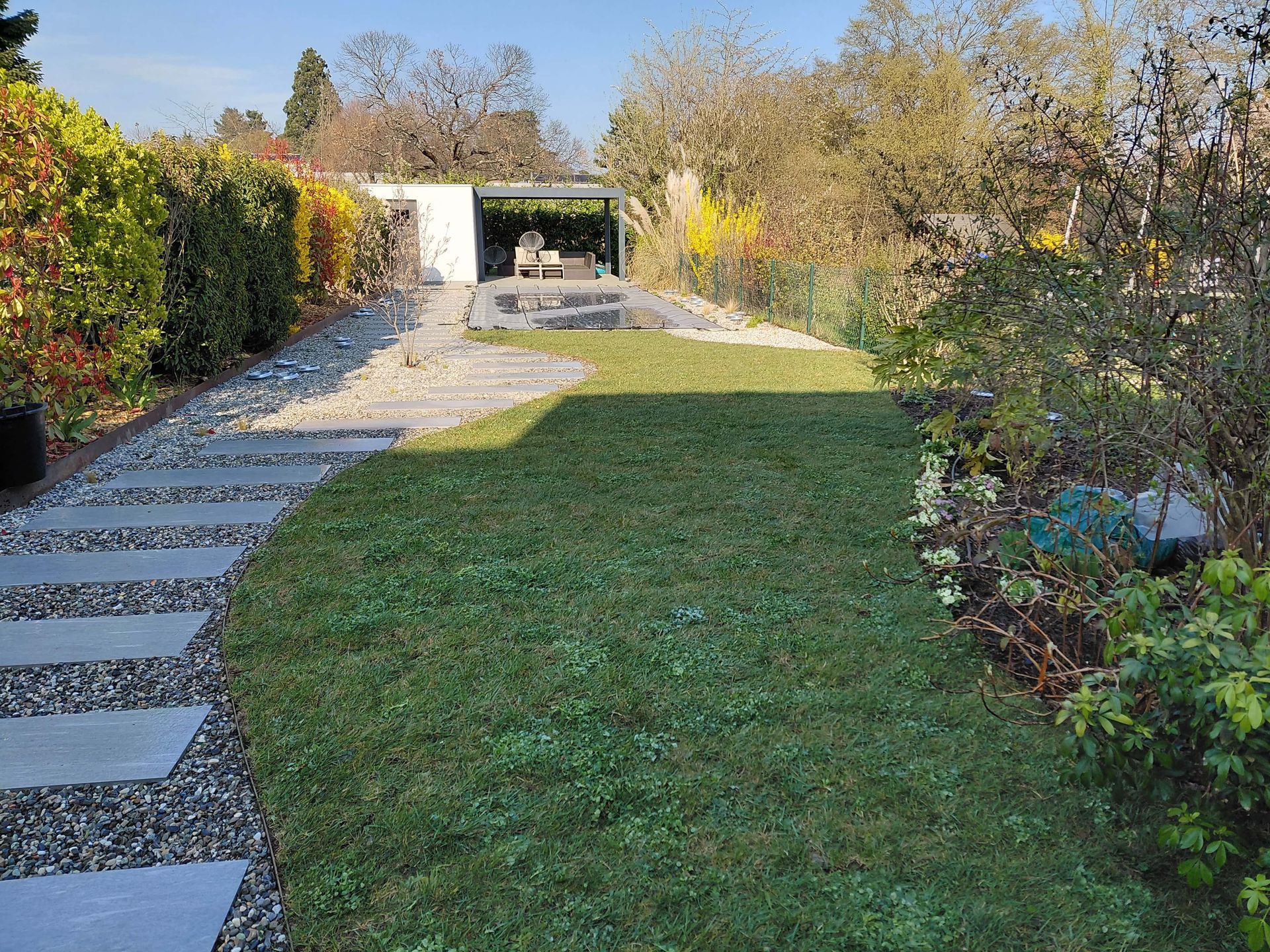 Allée de jardin avec dalles de pierre, pelouse verte et arbustes menant à une terrasse couverte.