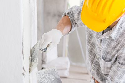 Ein Bauarbeiter streicht mit einem Pinsel eine weisse Wand. Er trägt einen gelben Schutzhelm und Handschuhe.