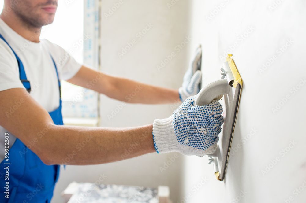 Ein Arbeiter in blauem Overall glättet mit einer Kelle Putz auf einer weissen Wand.