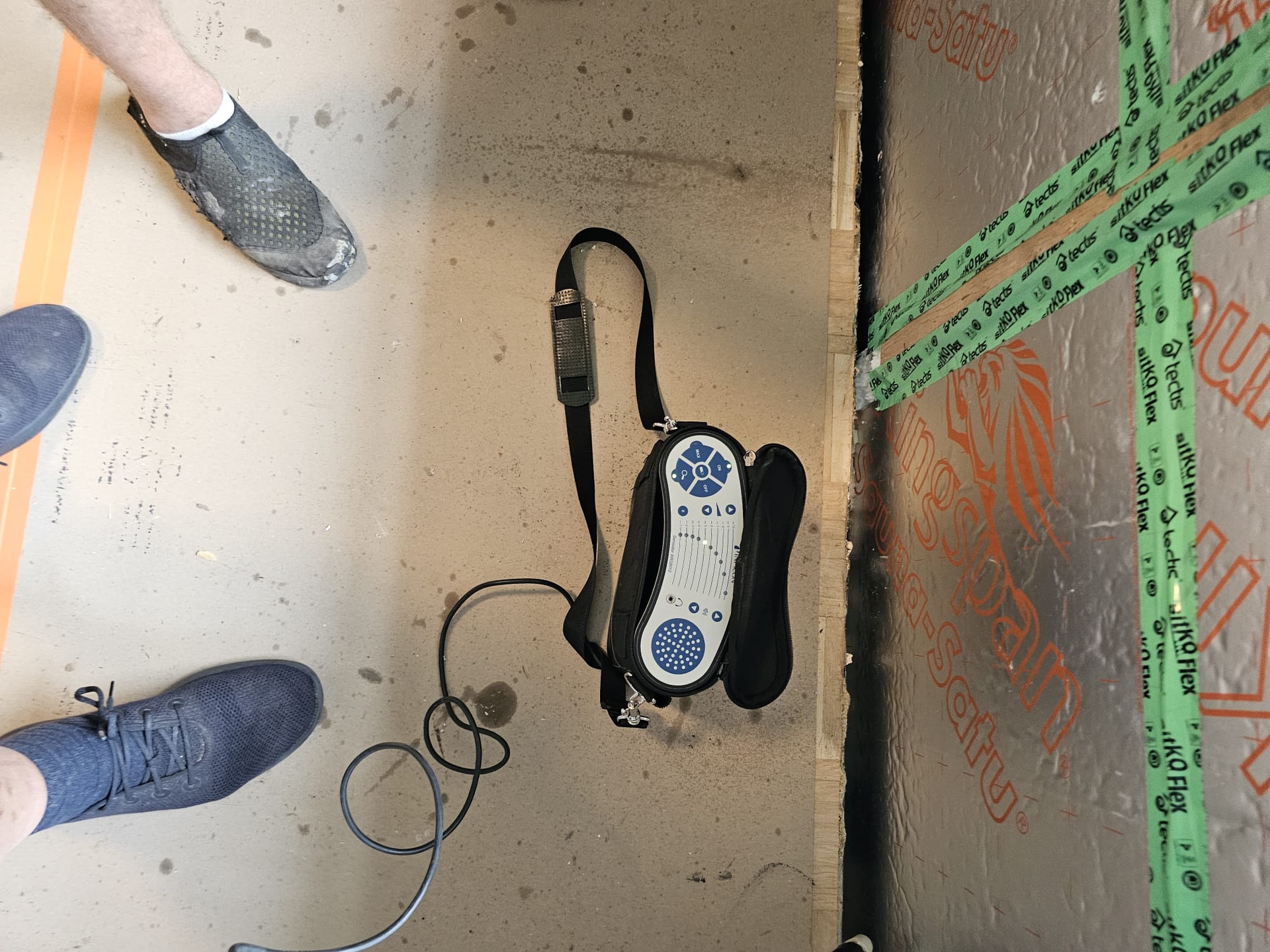 Jalat ympäröivät kosteusmittaria rakennustyömaalla. Laite on kytketty kaapeliin ja lepää lattialla.