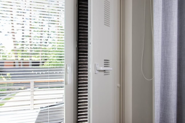 Valkoinen ikkuna, jossa kaihtimet ovat auki, valkoisen tuuletusaukon vieressä. Oikealla näkyy vaaleanharmaa verho.
