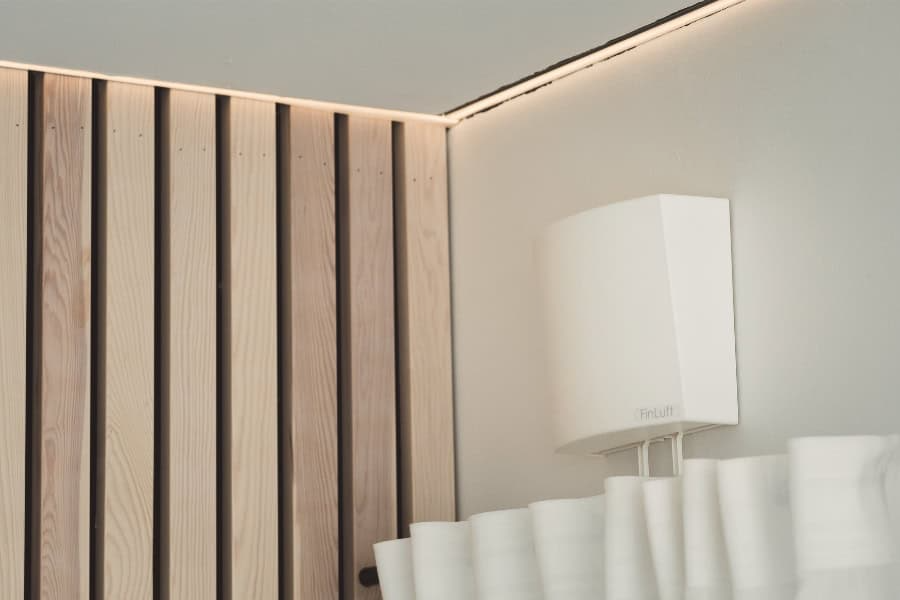 Valkoinen seinälle kiinnitetty laite johdoilla, lähellä puupaneeleita ja verhoa. Pehmeä valaistus korostaa nurkkaa.