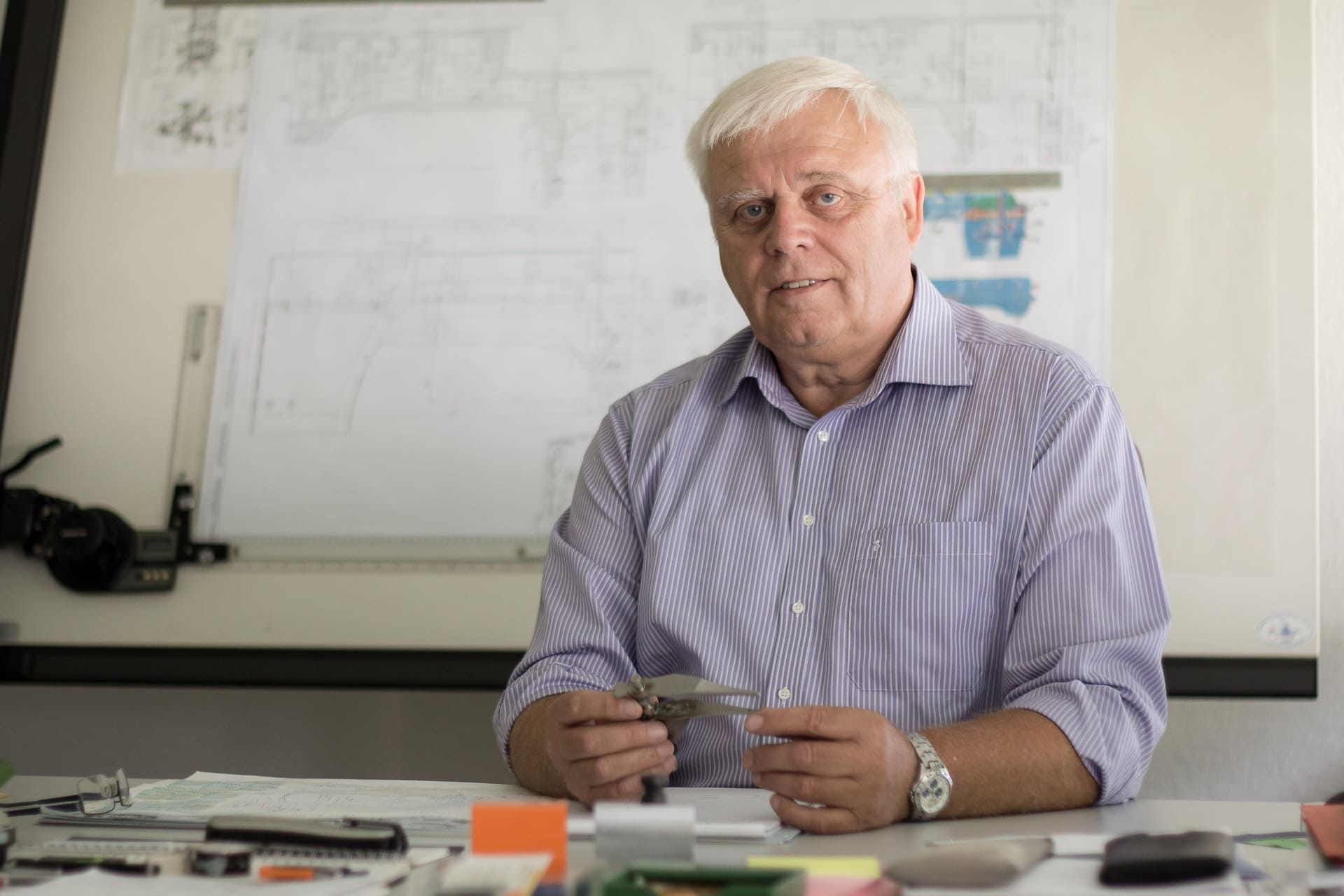 Ein Mann sitzt an einem Schreibtisch vor einer weißen Tafel und hält eine Lupe in der Hand.