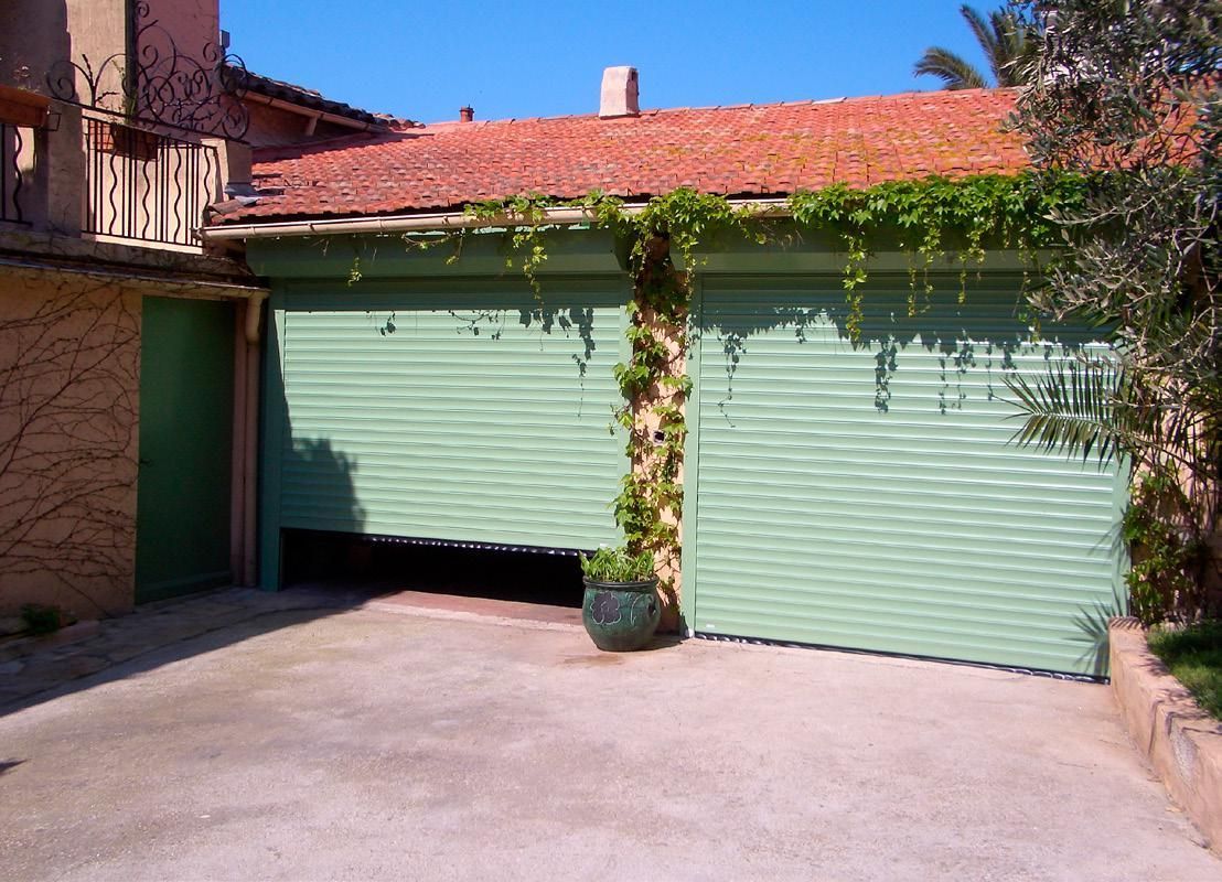 Des portes de garage vertes recouvertes de vigne, jouxtant un bâtiment en pierre et une allée menant à une cour.