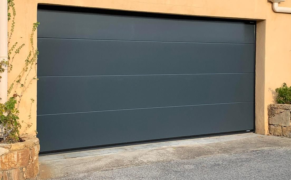 Porte de garage gris foncé fermée dans une allée bordée de murs en stuc beige et d'éléments en pierre.