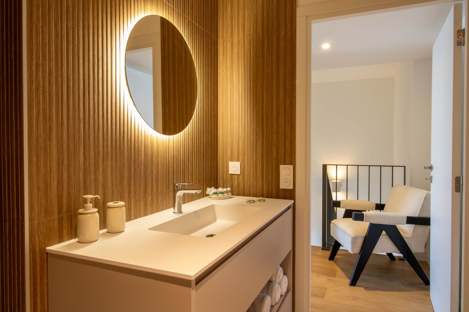Salle de bain avec mur en bois, miroir rond éclairé et lavabo blanc. Une porte ouverte mène à une pièce avec une chaise.