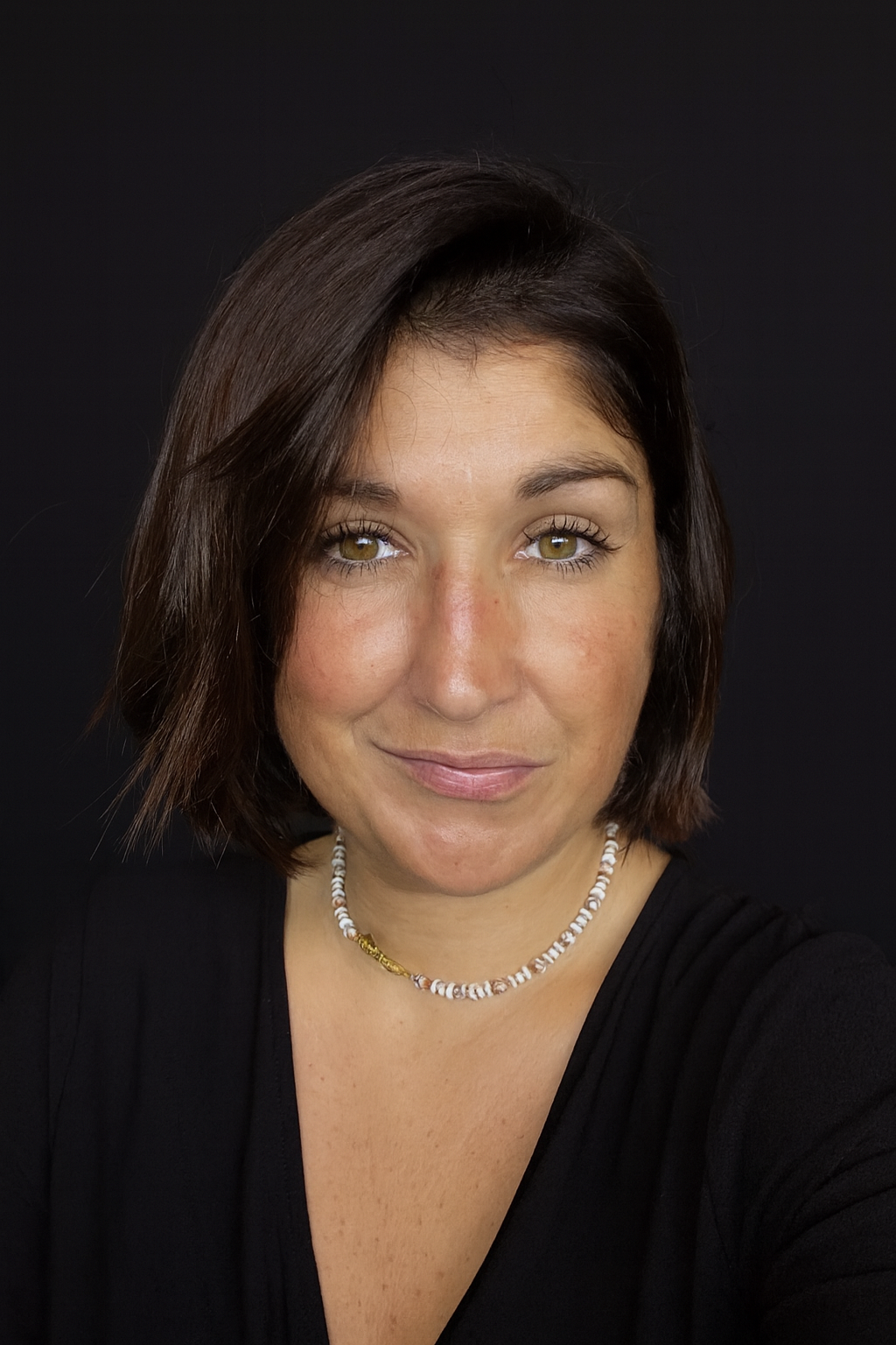 Femme aux cheveux noirs courts et aux yeux clairs portant un collier, souriant à la caméra, fond noir.