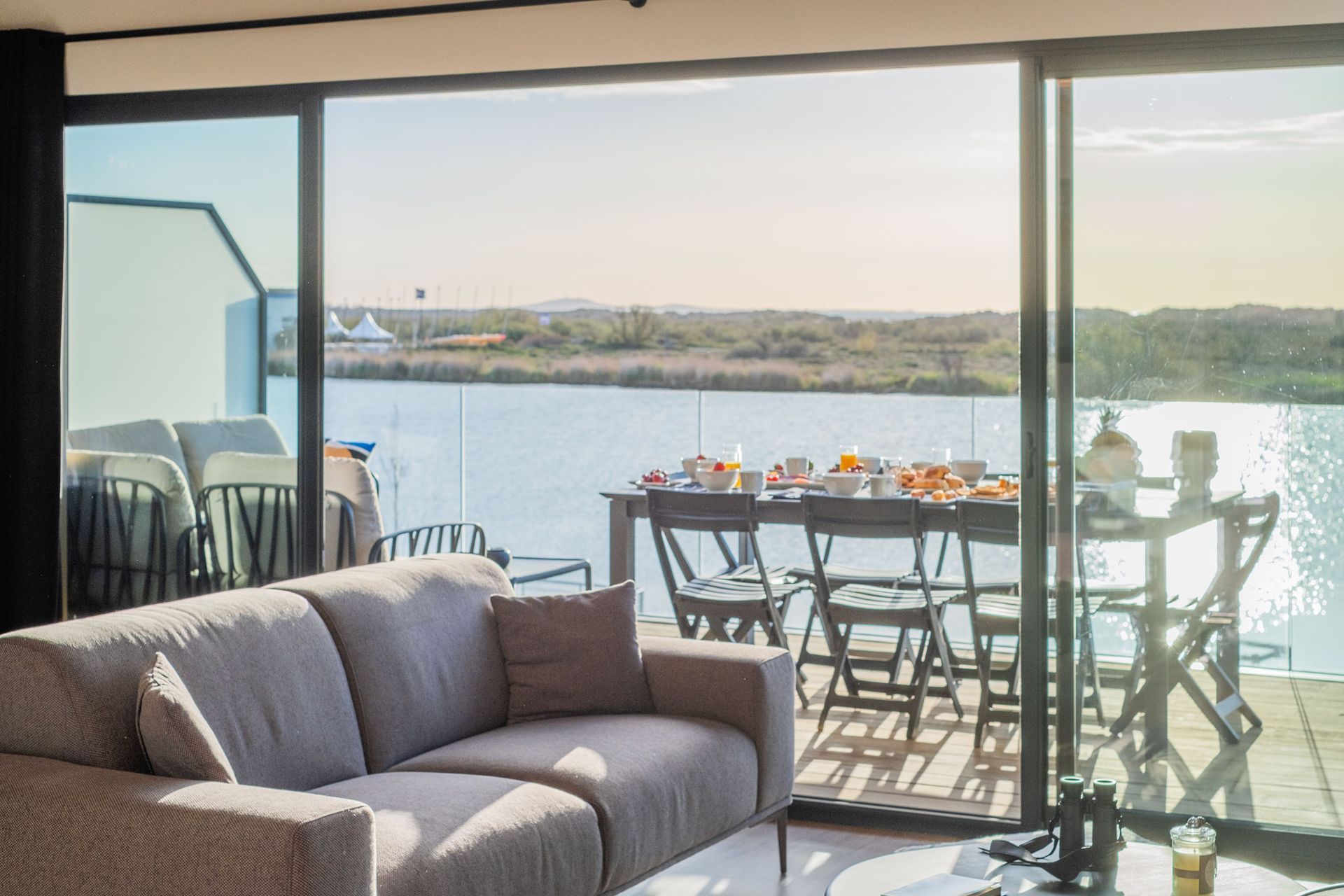 Salon avec vue sur l'eau. Canapé gris. Table de petit-déjeuner dressée sur une terrasse avec vue sur l'eau.