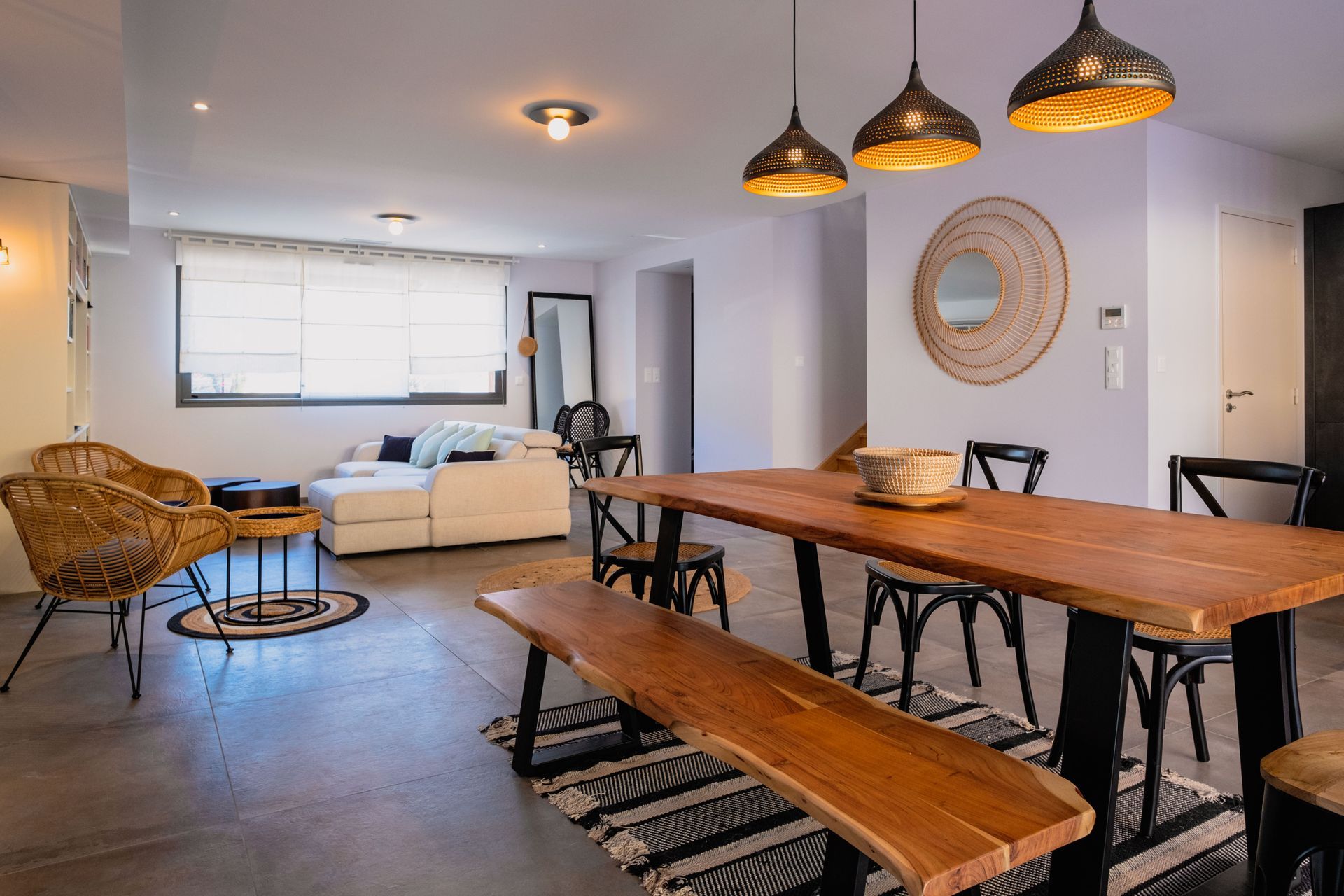 Séjour avec coin repas; table et banc en bois, chaises noires, canapé blanc, chaise tressée, suspensions.