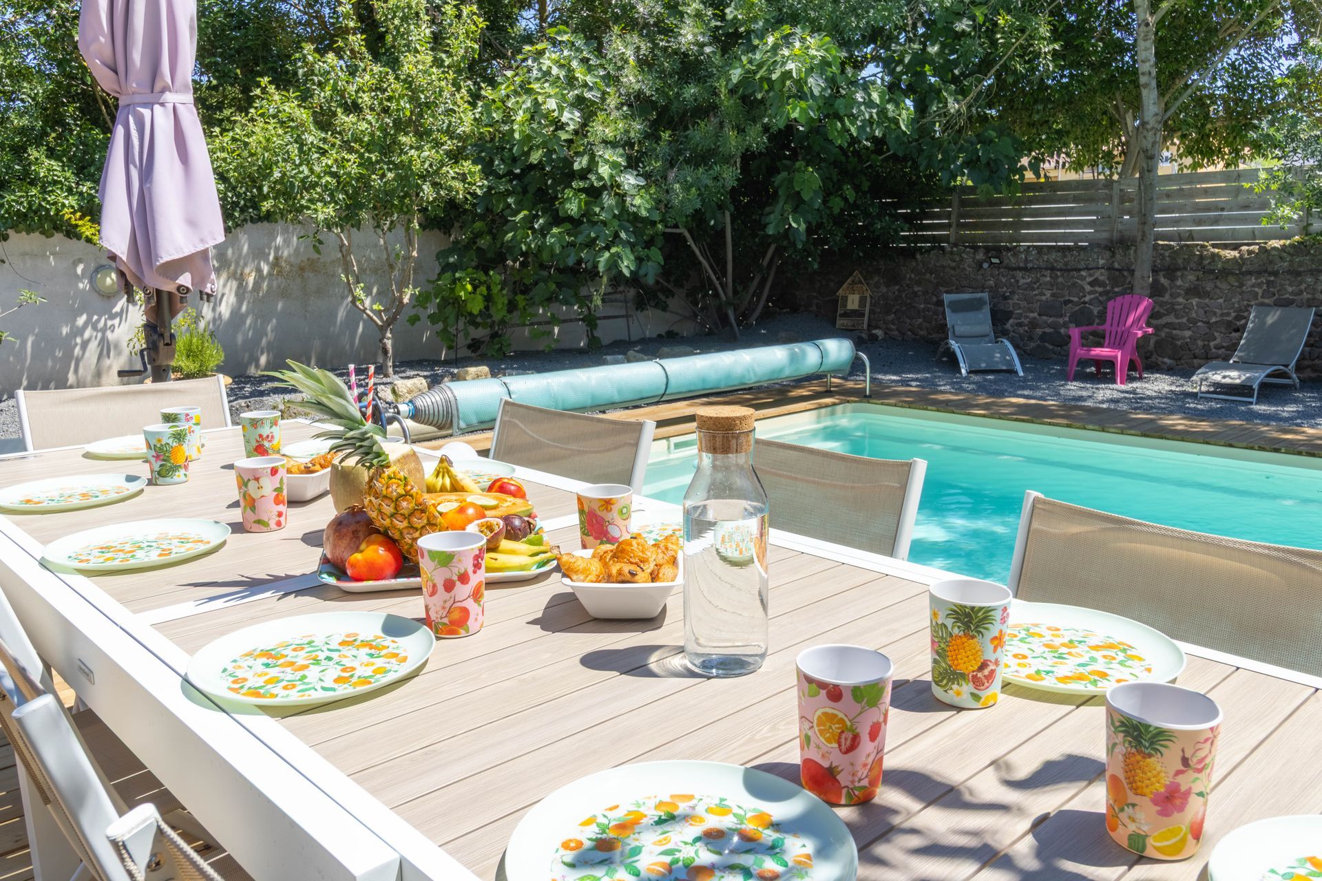 Table de piscine dressée pour un repas avec assiettes, fruits, boissons et chaises à côté d'une piscine.