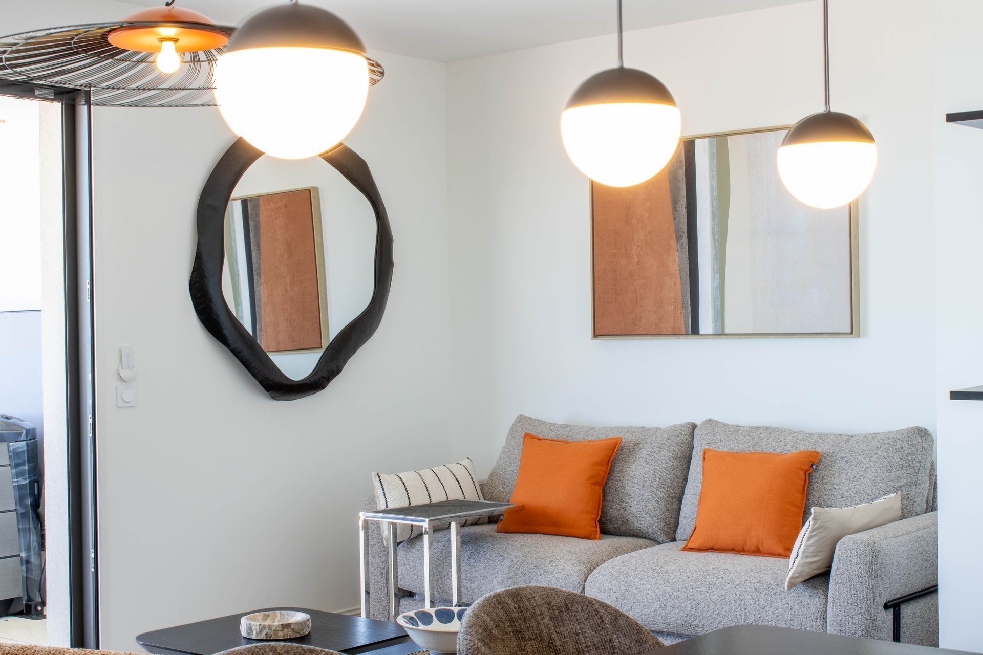 Salon avec canapé gris, coussins orange, miroir rond, œuvres d'art et suspensions globe.