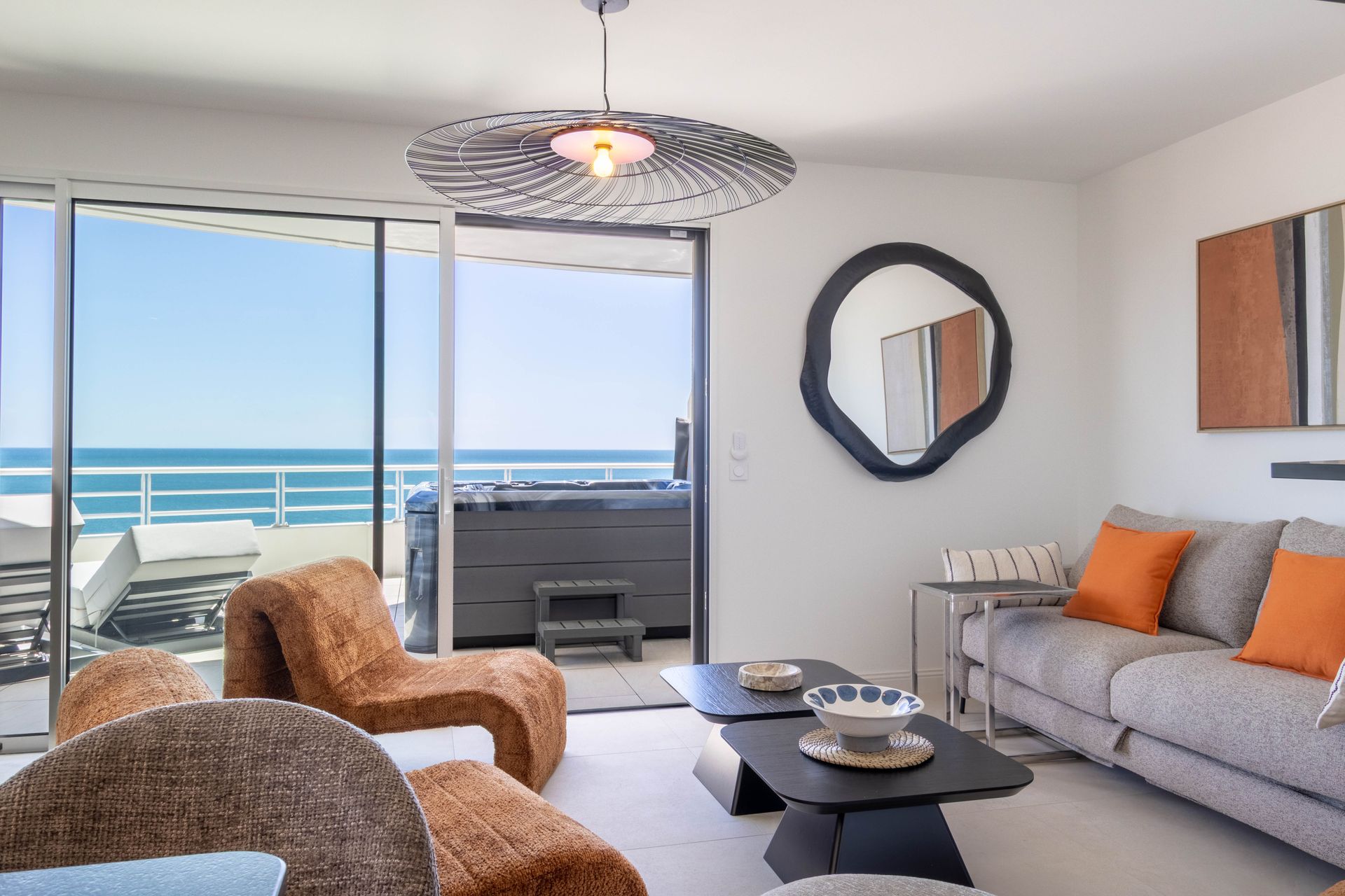 Salon avec vue sur l'océan, chaises marron, canapé gris, grand miroir et porte coulissante ouverte sur le bain à remous.