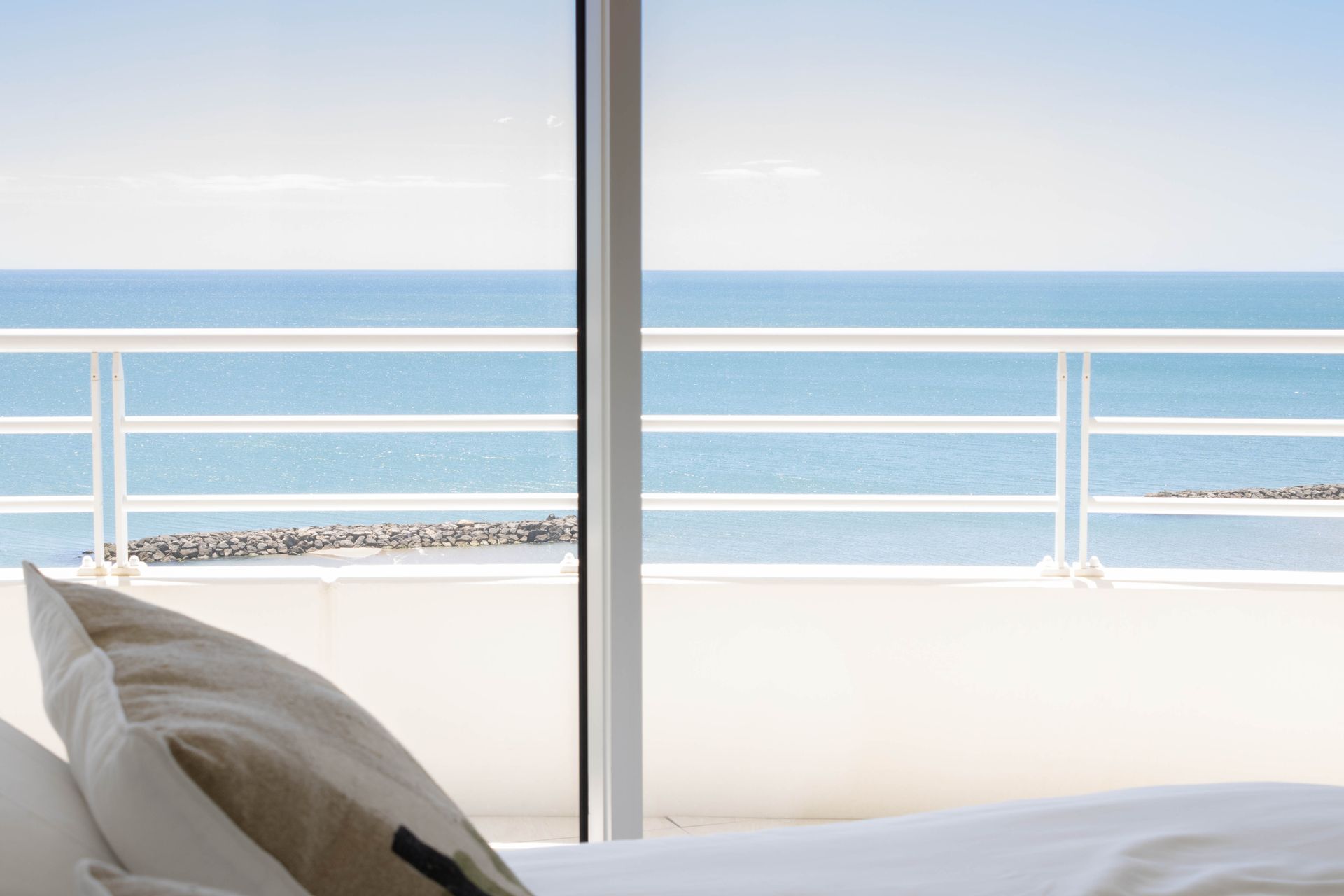 Chambre avec une grande fenêtre donnant sur l'océan et un balcon blanc. Un oreiller repose sur un lit blanc.