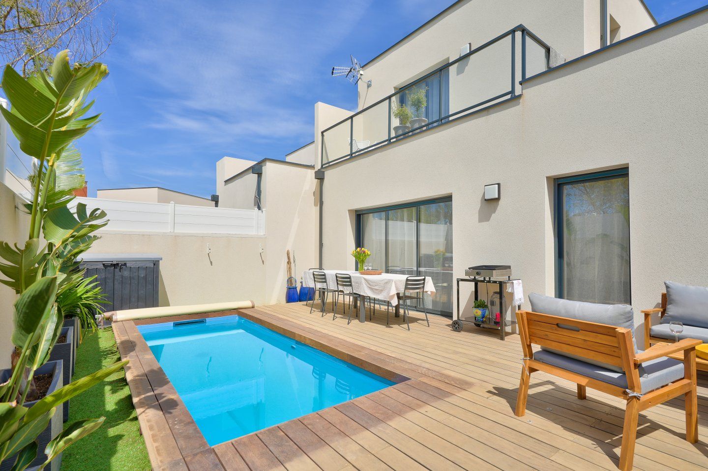 Jardin avec petite piscine, terrasse, coin repas et bâtiment blanc de deux étages sous un ciel bleu.