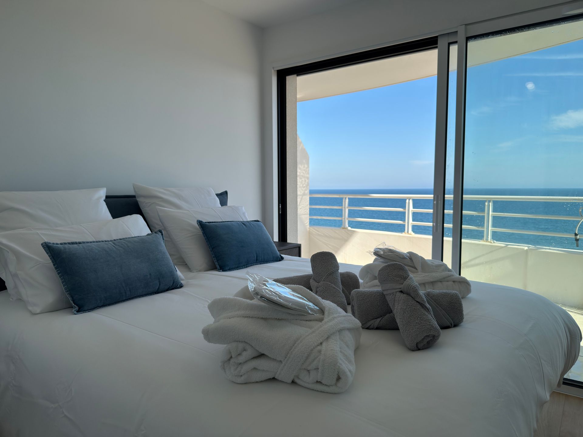 Chambre avec un lit, des oreillers bleus et vue sur l'océan à travers une porte coulissante en verre.