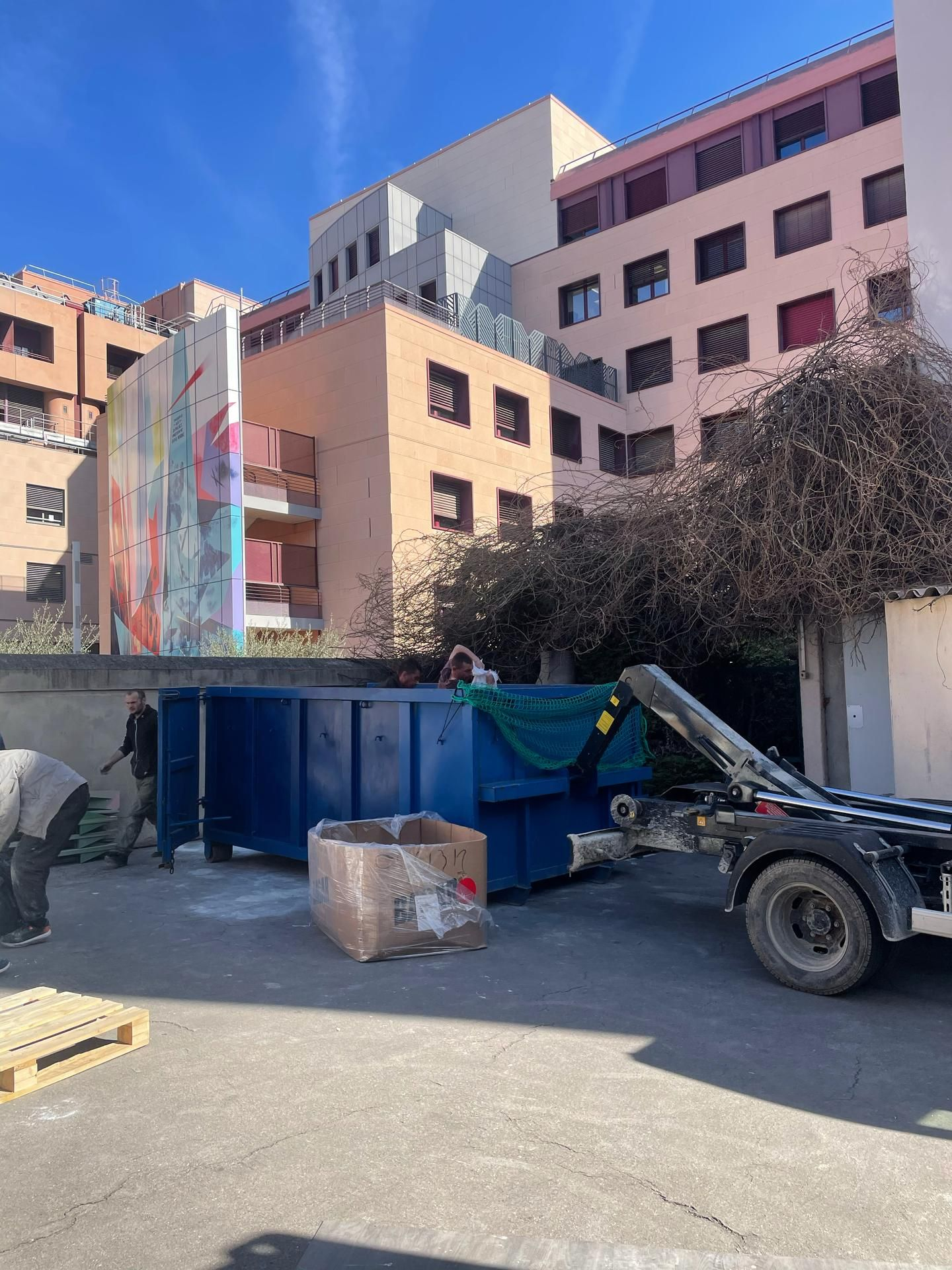 Des ouvriers chargent une benne bleue devant des bâtiments