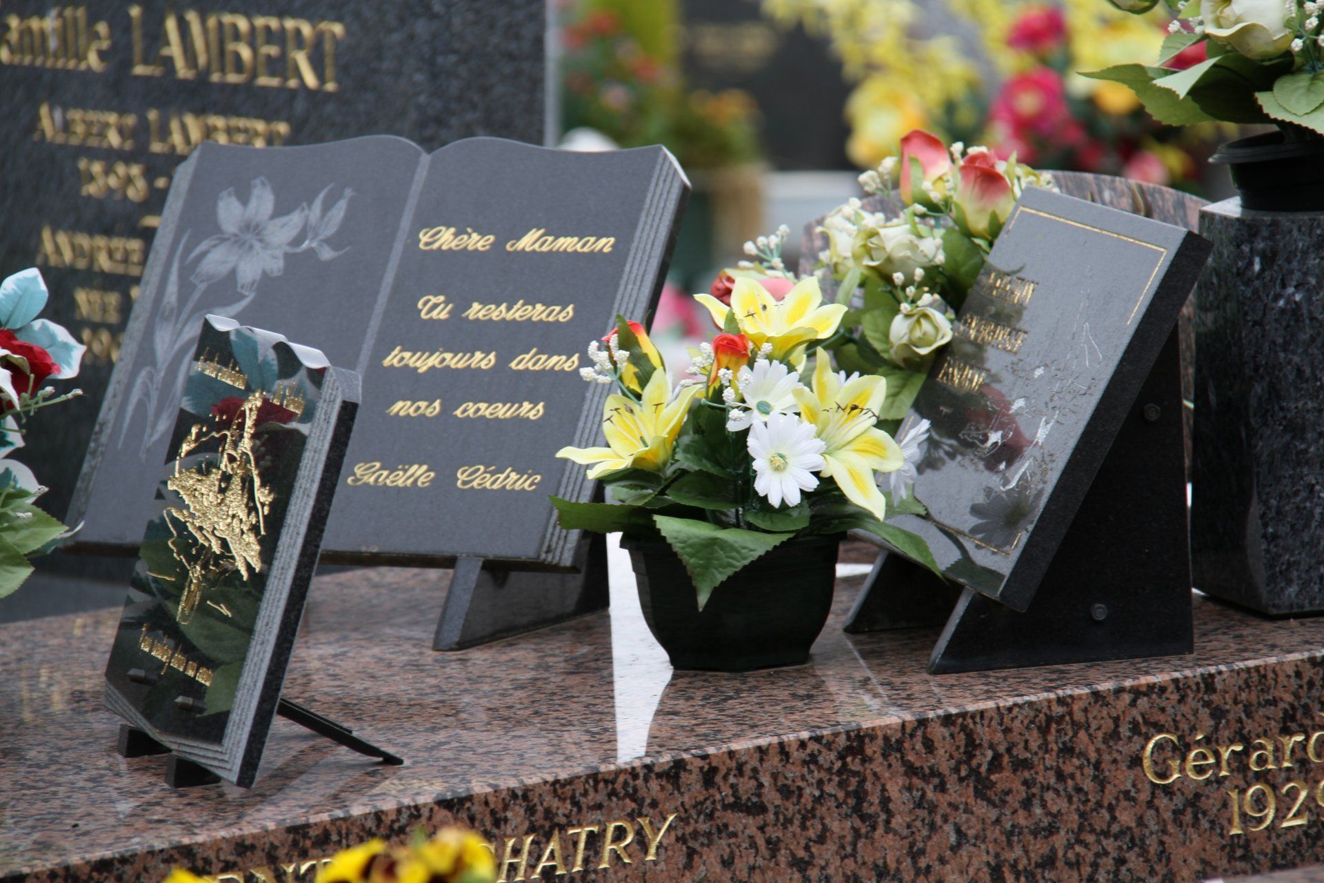 Scène de cimetière avec pierres tombales et compositions florales. Une pierre tombale en forme de livre porte une inscription.