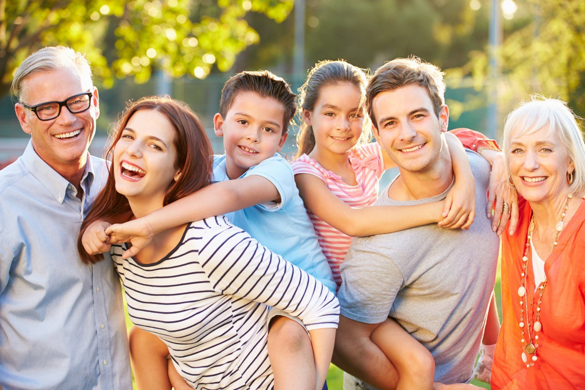 Une famille souriante dans un parc, avec enfants, parents et grands-parents. Lumière du soleil et fond vert.